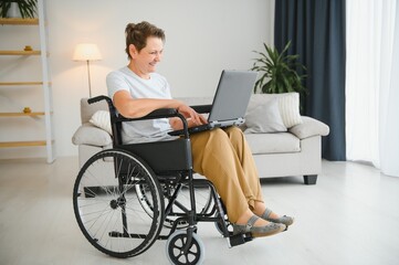 Middle age grey-haired disabled woman having video call sitting on wheelchair at home.