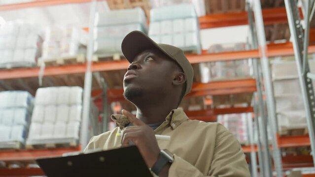 Low angle shot of African American warehouse manager examining shelves and taking notes on clipboard while doing inventory at work