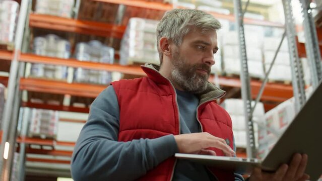 Low angle shot of middle aged manager standing in warehouse and typing on laptop during workday