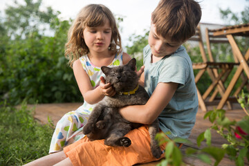 two children are holding and petting a cat in nature near the house
