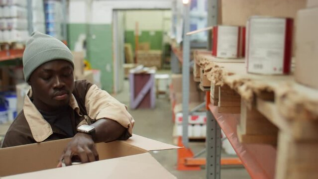 African American male worker taking paint cans from shelf and putting them in cardboard box while picking order in warehouse