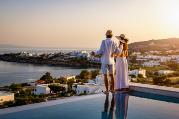 Happy couple on vacation time enjoys the summer sunset over the Aegean Sea by the swimming pool with an aperitif drink - 516279874