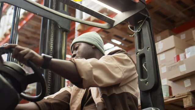 Low angle shot of African American warehouse worker leaning back on driver seat of forklift, pulling hat on eyes and falling asleep with arms crossed while taking break during workday