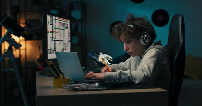 A boy sits in room in the evening in front of computer screen and laptop, working on a school project, drawing in a notebook with a pencil, planning the look of his work, thinking.