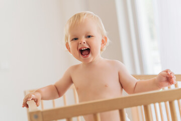 Portrait of funny 11 month old blond baby standing in bed holding onto bumpers shouting with...