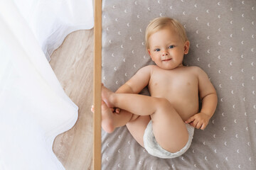 Upper view of sweet cute blue-eyed baby in diaper lying on his back in crib looking at camera, playing with foot putting legs up. Childcare concept. Happy carefree childhood. Copy space - obrazy, fototapety, plakaty