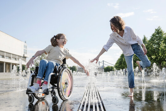 Woman and daughter with disability playing along fountain at park on sunny day