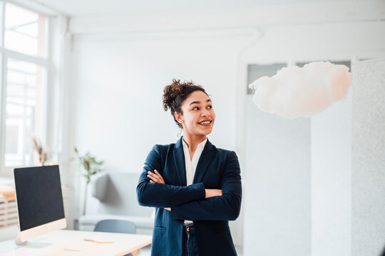 Happy young businesswoman with arms crossed looking at cloud levitating in office