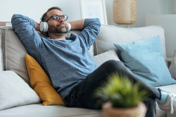 Smiling man listening music through headphones relaxing on sofa at home