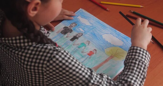 Schoolgirl in a plaid dress with brown hair braided in pigtails sitting at a table in a classroom and drawing a picture of family in an album using color pencils.  