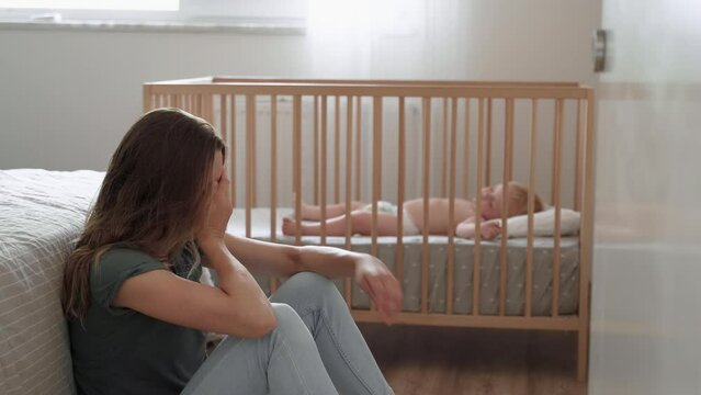 Young mother suffering postnatal depression sitting on floor next to baby napping in bed, resting head on knees, crying, feeling desperate in need of professional psychological support