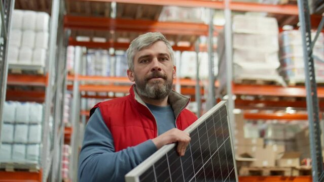 Portrait of middle aged man holding solar panel and posing for camera at work in warehouse