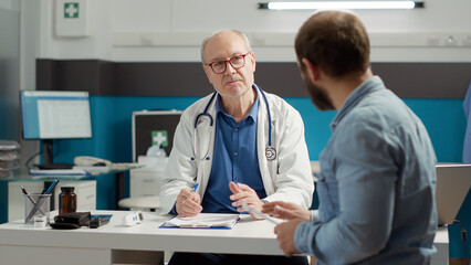 Male physician writing checkup visit report on medical files, preparing prescription medicine to...