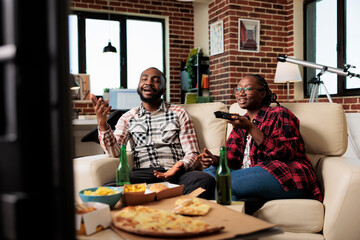 Happy girlfriend and boyfriend switching tv channels and eating takeaway meal from fast food delivery at home. Watching movie on television program and enjoying takeout food with beer bottles.