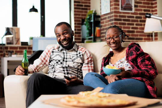 Smiling Couple Watching Film On Television And Eating Takeaway Meal From Delivery, Having Fun Together. Enjoying Fast Food Takeout And Bottles Of Beer While They Watch Movie On Tv.