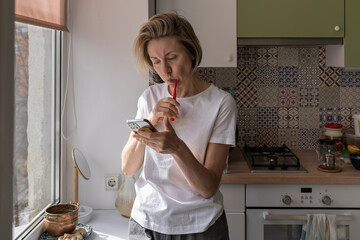 Busy scandinavian woman using mobile smartphone, talking in video chat with friend, chatting in social networks, watching video check mobile application while brushing teeth next to the window at home