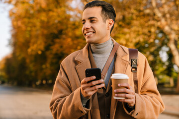 Young man using cellphone and drinking coffee in autumn park