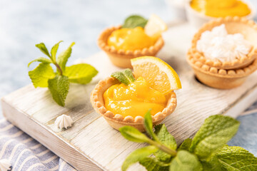 Shortbread tartlet filled with lemon curd, mint and lemon slices and mini meringue on wooden board, blue background. - obrazy, fototapety, plakaty