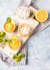Shortbread tartlet filled with lemon curd, mint and lemon slices and mini meringue on wooden board, blue background. Top view.