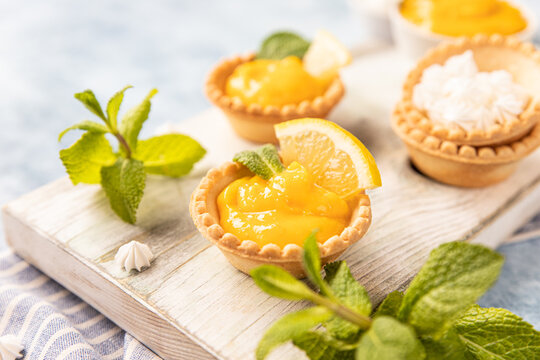 Shortbread tartlet filled with lemon curd, mint and lemon slices and mini meringue on wooden board, blue background.