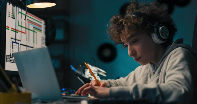 A young boy sits at his desk with wireless headphones in front of a computer taking notes with a pencil, drawing a design, passionate about architecture.