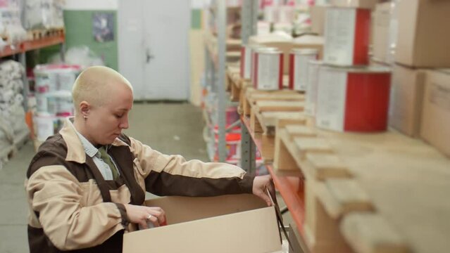 Female worker packing paint cans from shelf in cardboard box and then walking away while picking order in warehouse