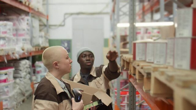 Caucasian woman scanning barcodes on paint cans as her African American male colleague packing them in cardboard box while picking order together in warehouse