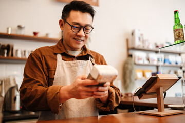 Adult happy asian man holding payment terminal at counter in cafe