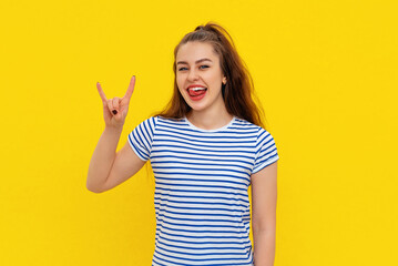 Emotional brunette girl makes rock n roll gesture, exclaims, feels overjoyed, enjoys music theme party, standing in white-blue striped t shirt over yellow background. Body language concept