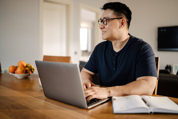 Brunette adult asian man smiling and using laptop computer at home