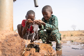 Two happy boys playing with fresh water from a village tap in an African steppe region; desertification progress and water scarcity concept