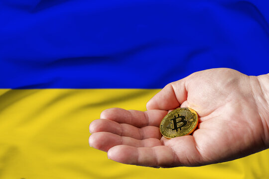 Golden bitcoin coin in man's hand, Ukrainian flag in the background