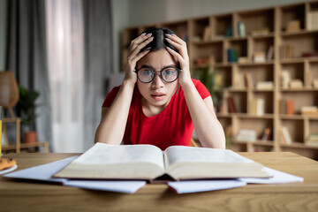 Disappointed shocked adolescence asian female student in glasses reads book, hold head with hand