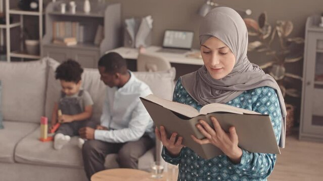 Medium slowmo portrait of young Muslim woman in grey hijab reading Quran at home while her African American husband playing with their little son on sofa in background