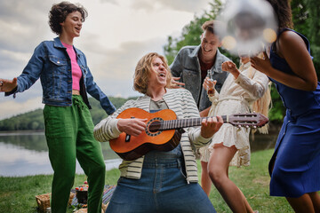 Fototapeta premium Group of young friends having fun on picnic near a lake, laughing, dancing and playing guitar.
