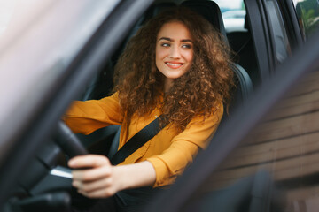 Young woman driving a car in the city. Portrait of a beautiful woman in a car, looking out of the window and smiling. - obrazy, fototapety, plakaty