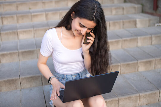 the girl is sitting on the stairs, laptop on her lap, talking on the phone and laughing