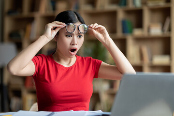 Sad shocked adolescent chinese girl with open mouth takes off glasses looks at computer in room...