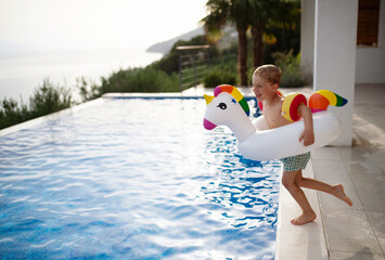 Happy kid having fun when jumping to swimming pool with inflatable ring unicorn. Summer outdoor...