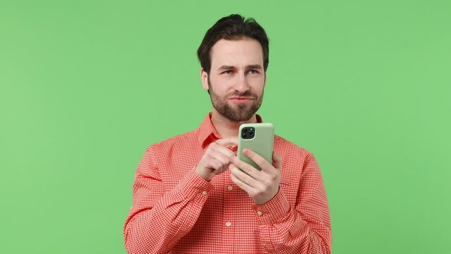 Dreamful pensive minded excited young brunet bearded man 20s years old wears pink shirt hold using mobile cell phone typing browsing chatting send sms isolated plain green background studio portrait