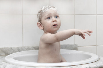 Naklejka premium A small child sits in a ceramic sink in the bathroom. Baby bath.