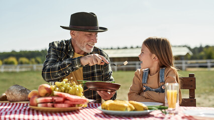 Grandfather and girl have lunch at table on farm