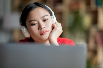 Sad boring adolescent asian lady student in headphones watching video lesson on computer in room...