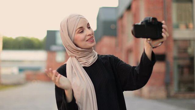 Young female muslim blogger in trendy clothes and hijab walks on city street, records herself on video holding camera in hand. Arab girl takes pictures of herself on camera. Lifestyle selfie backlight