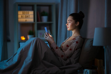 technology, bedtime and rest concept - teenage girl in pajamas with smartphone sitting in bed at...