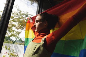 Image of caucasian non-binary trans woman holding rainbow flag and looking outside window