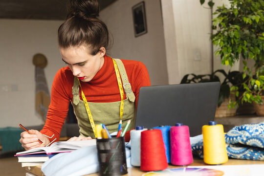 Image of caucasian non-binary trans woman with laptop and notes working in sewing workshop