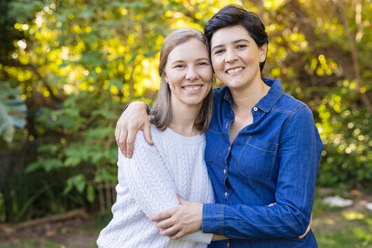 Portrait of happy caucasian mid adult lesbian couple with arms around standing against trees in yard