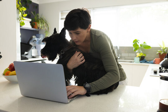 Caucasian mid adult lesbian woman carrying black scottish terrier and using laptop on kitchen island