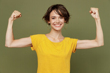 Young strong sporty fitness woman happy woman she 20s wear yellow t-shirt showing biceps muscles on hand demonstrating strength power isolated on plain olive green khaki background studio portrait